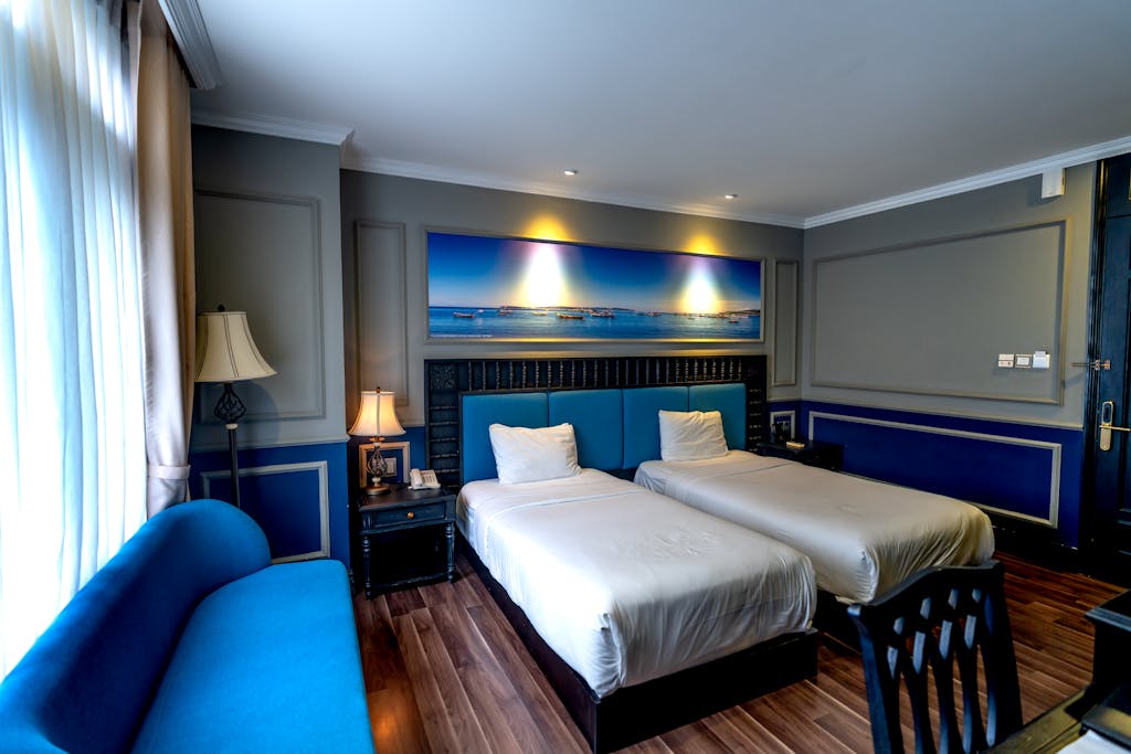 Stylish hotel room featuring blue decor, twin beds, and modern furnishings with ample natural light.