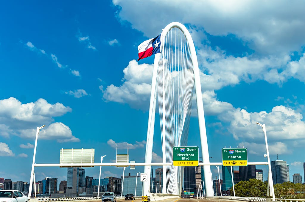 Iconic view of Margaret Hunt Hill Bridge with Texas flag and Dallas skyline under a blue sky.